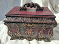 May include: A burgundy decorative box with elaborate carvings and a handle. The rectangular box features a detailed, raised design with gold and green accents. The lid is flat and the box rests on small feet.