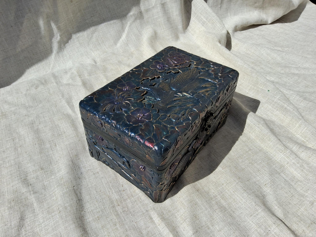 May include: A rectangular, dark blue box with detailed floral and bird carvings. The box has a metallic sheen with purple and bronze highlights. The carvings are intricate and cover the top and sides, suggesting a decorative or storage item.